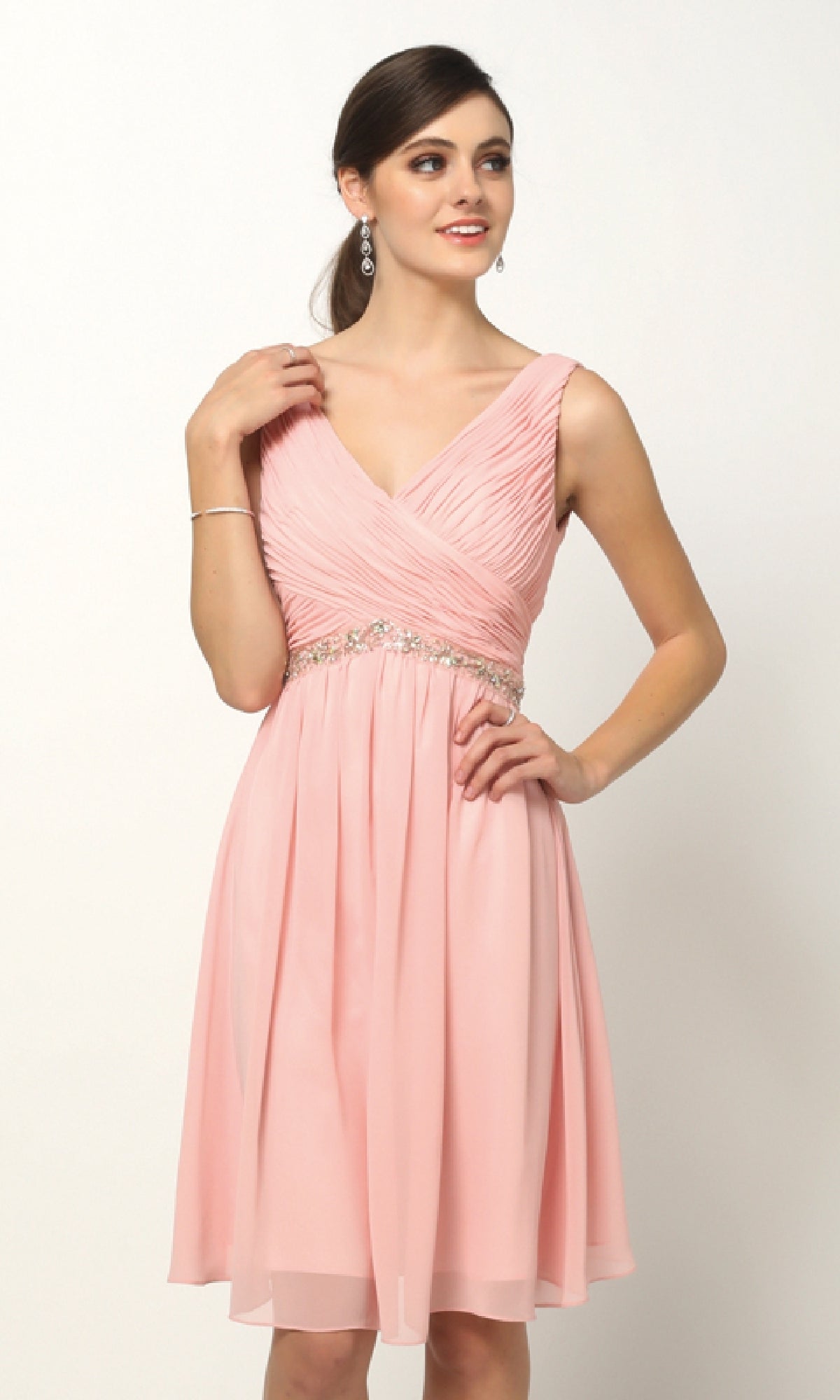 A woman models the Juliet 786 short semi-formal dress, featuring an A-line silhouette in pink with a V-neckline and beaded empire waist, standing against a plain white background.